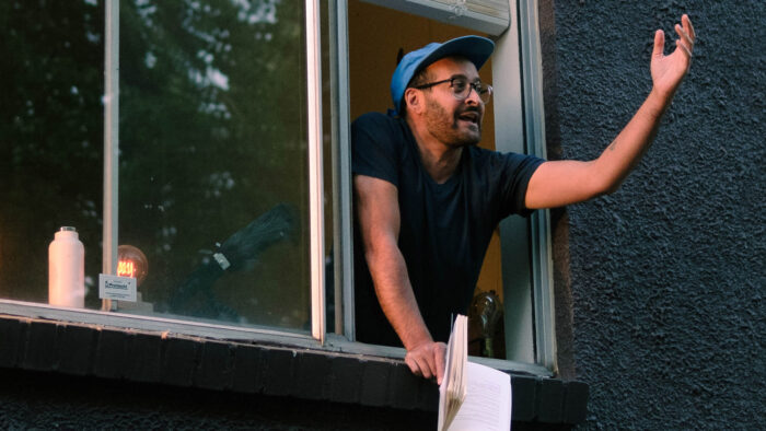 A man with a blue hat, eyeglasses, and a black t-shirt is holding a book and his hand out a window, like he's speaking to people outside