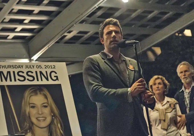 A man stands at a microphone in front of a photo of a blond, white woman with MISSING above her photo