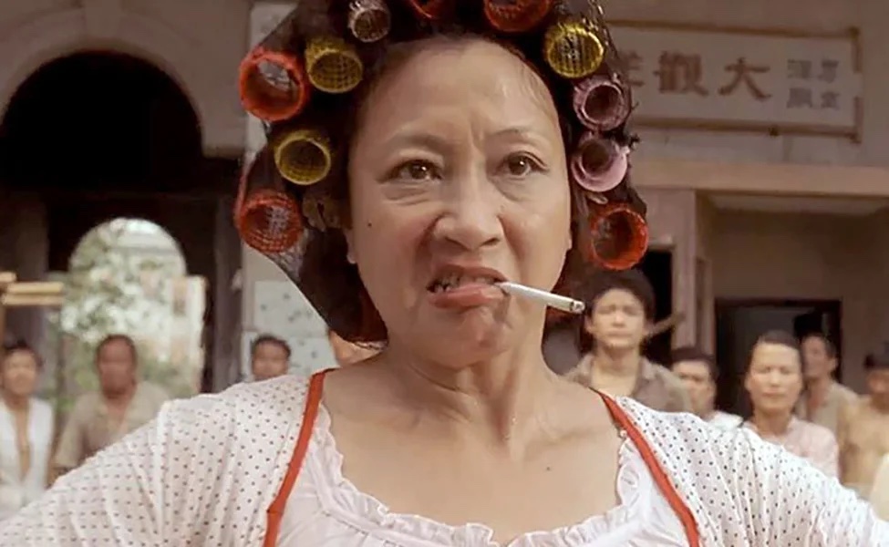 An angry-looking woman in a nightgown with curlers in her hair and a cigarette hanging from her mouth stands in front of a crowd of people