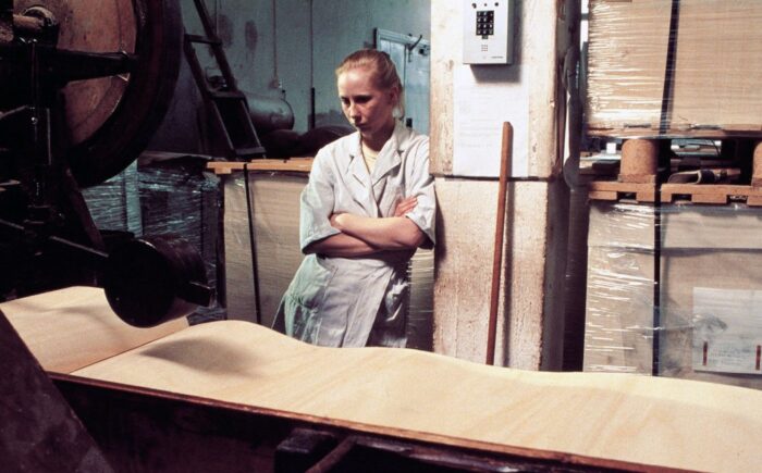 A young blonde woman stands in a factory, leaning against the wall with her arms crossed