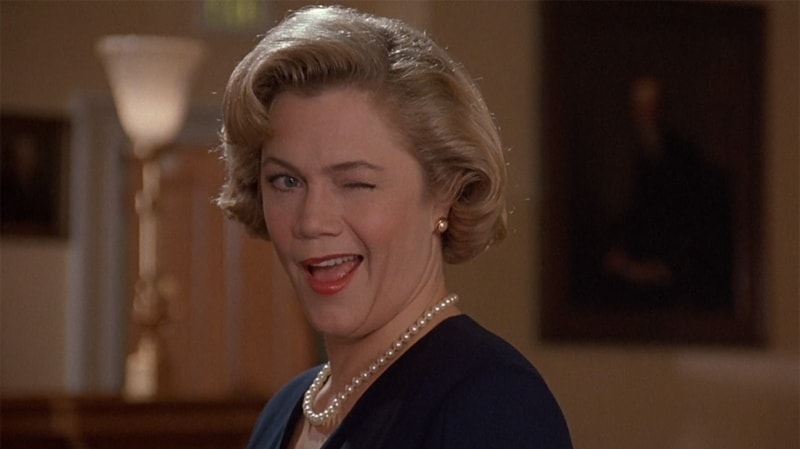 A middle-aged woman with a blond coiffed bob and a pearl necklace winks at the camera