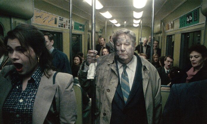 A subway car full of sad and scared looking people. A middle-aged man stands in the middle in a suit and trench coat, with dirt on his face and clothes