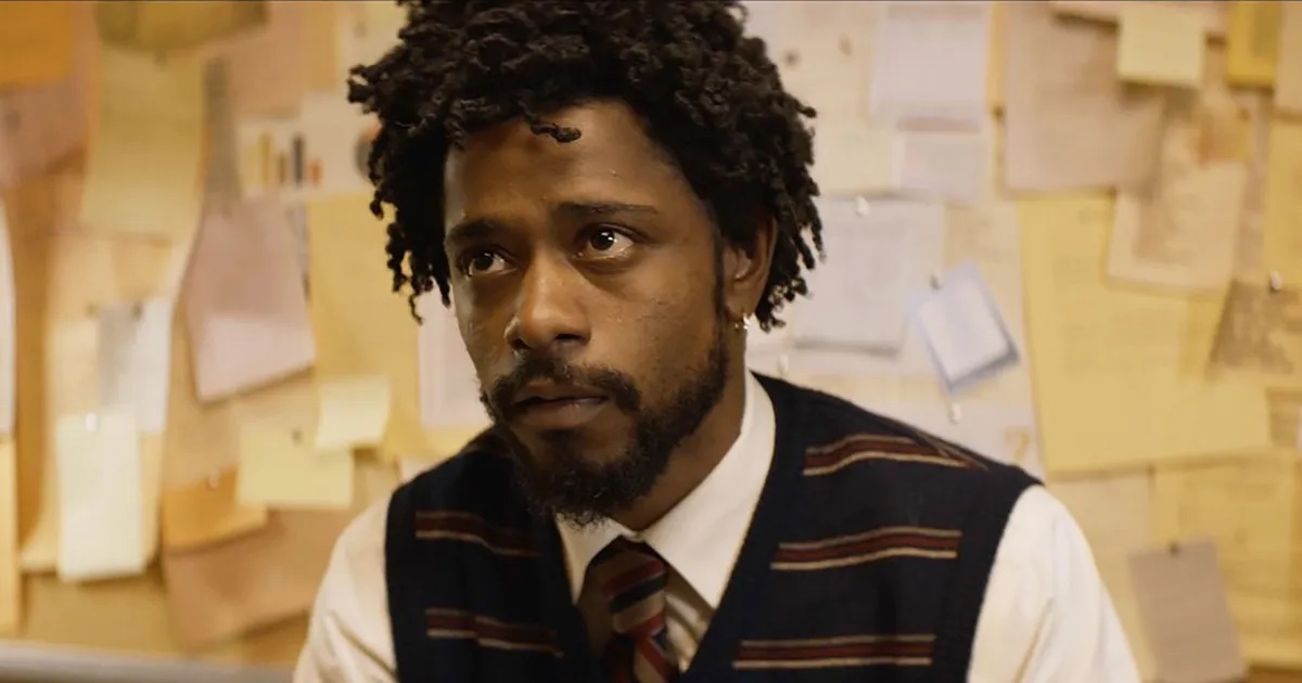 A young Black man with short locs, a white button-down shirt, tie, and a sweater vest, sits in front of a wall of post-it notes