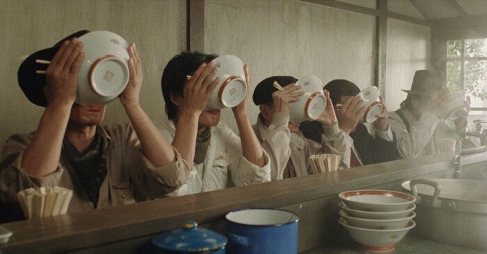 A group of people sitting at the bar of a ramen shop with bowls tipped up in front of their faces