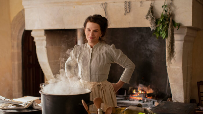 Film still of a woman in a late 1800's dress standing in a kitchen in front of a steaming pot