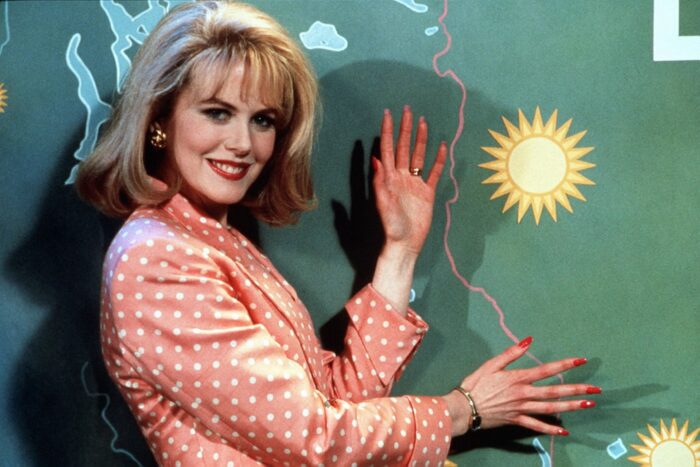 A woman with a blond bob and an orange checkered suit jacket stands in front of a weather map