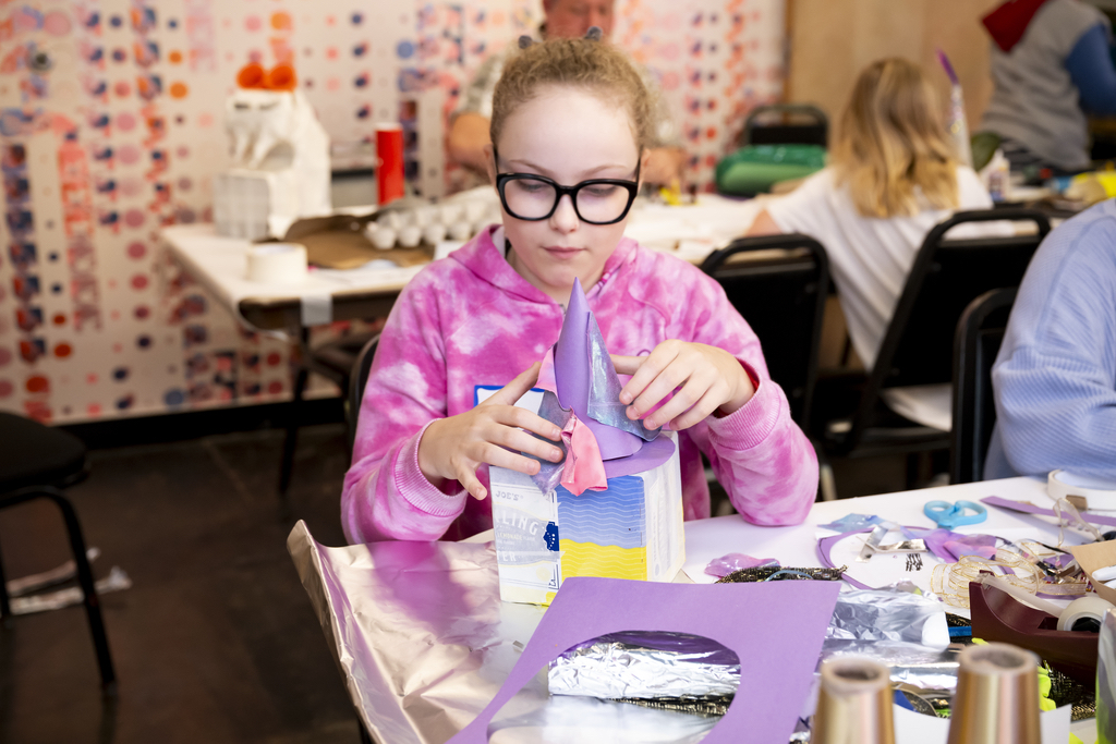 A girl with glasses and a pink hoodie on sitting at a table full of arts and craft supplies