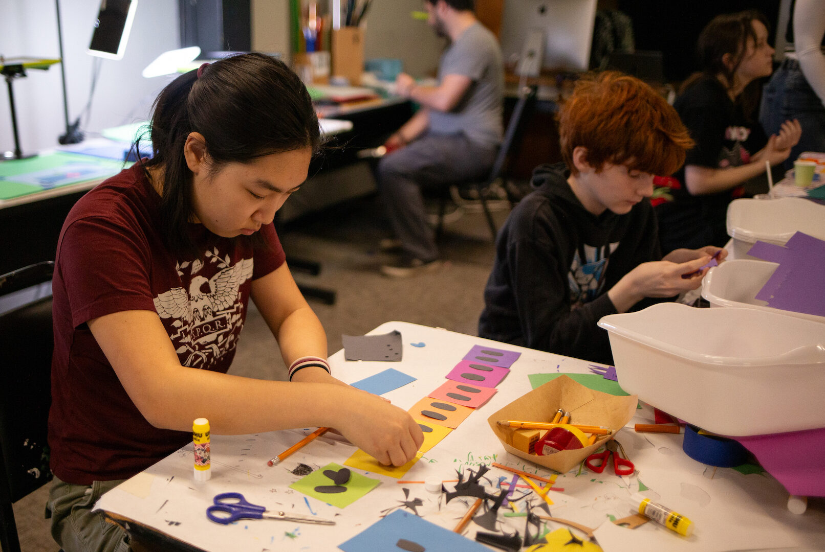 Photo of a young woman sitting at a table and concentrating on art that she's making