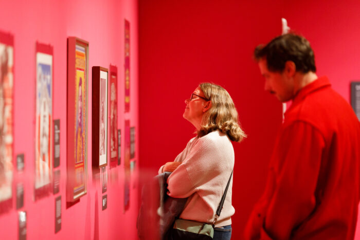 A woman with eyeglasses and a bob haircut and a man with a red jacket and a mustache looking at a bright pink wall with framed posters