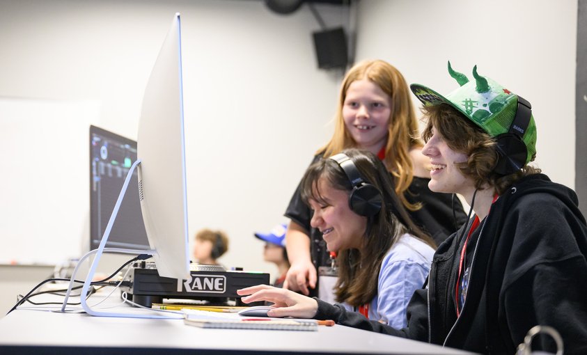 Photo of three kids looking at a computer and smiling. Two of them are wearing headphones.