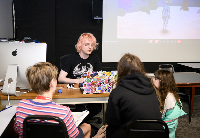 Photo of an adult with pink hair and eyeglasses, with a screen behind them, sitting across a desk from three kids