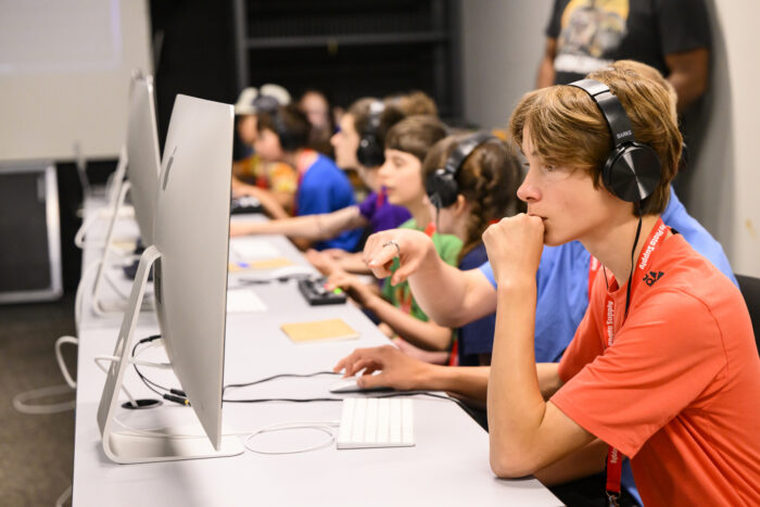 Photo of a line of middle-school aged kids wearing headphones, sitting at a table in front of computers