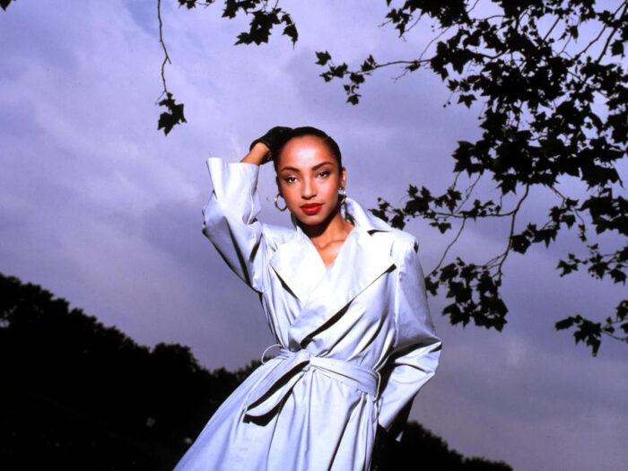 A photo of a Black woman (Sade) with slicked back hair, wearing a white trenchcoat, in front of a purple sky