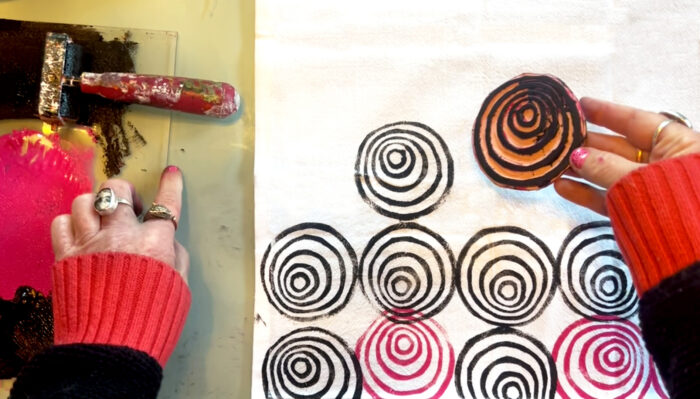 A photo of two hands, wearing a red and black sweater and rings, holding a spiral block print over fabric where the print is repeated in red and black. There's a roller and black ink next to one hand.