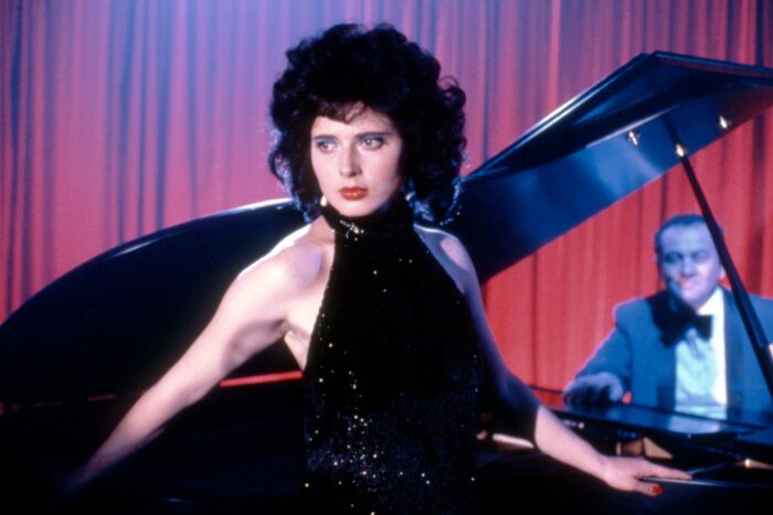 A woman with curly dark hair and a sequined black dress standing in front of a grand piano, a seated man in a tuxedo, and a red curtain