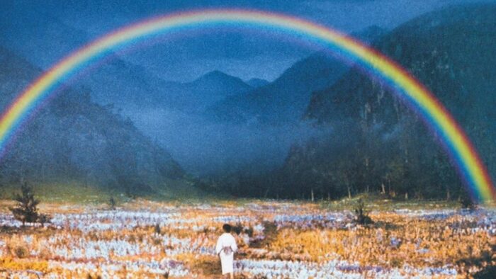 Photo of a valley with mountains and orange and white flowers and a rainbow. A child wearing a white kimono stands in the foreground.