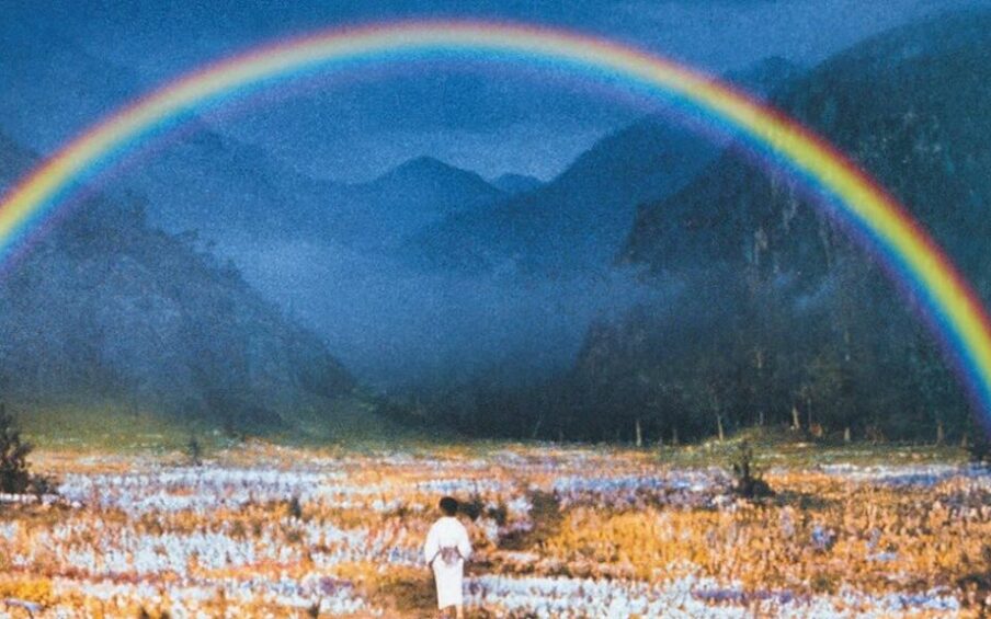 Photo of a valley with mountains and orange and white flowers and a rainbow. A child wearing a white kimono stands in the foreground.