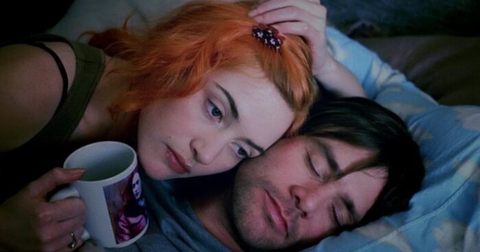 A woman with orange hair laying her head on top of a sleeping man's head. She is holding a mug.