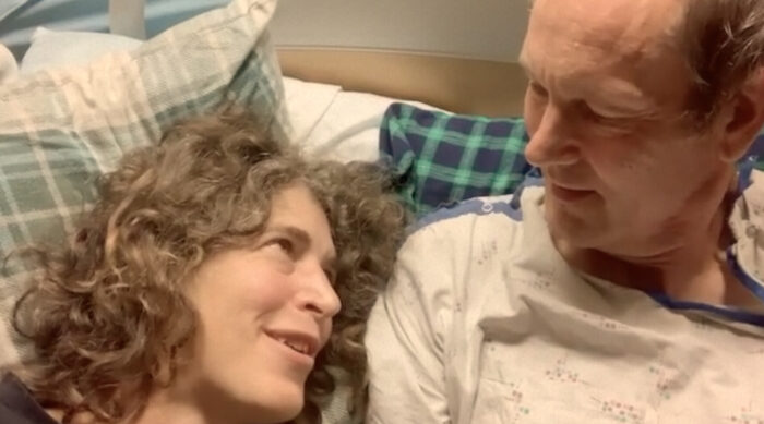 A photo of a woman with blond curly hair laying in bed with a man in a hospital gown