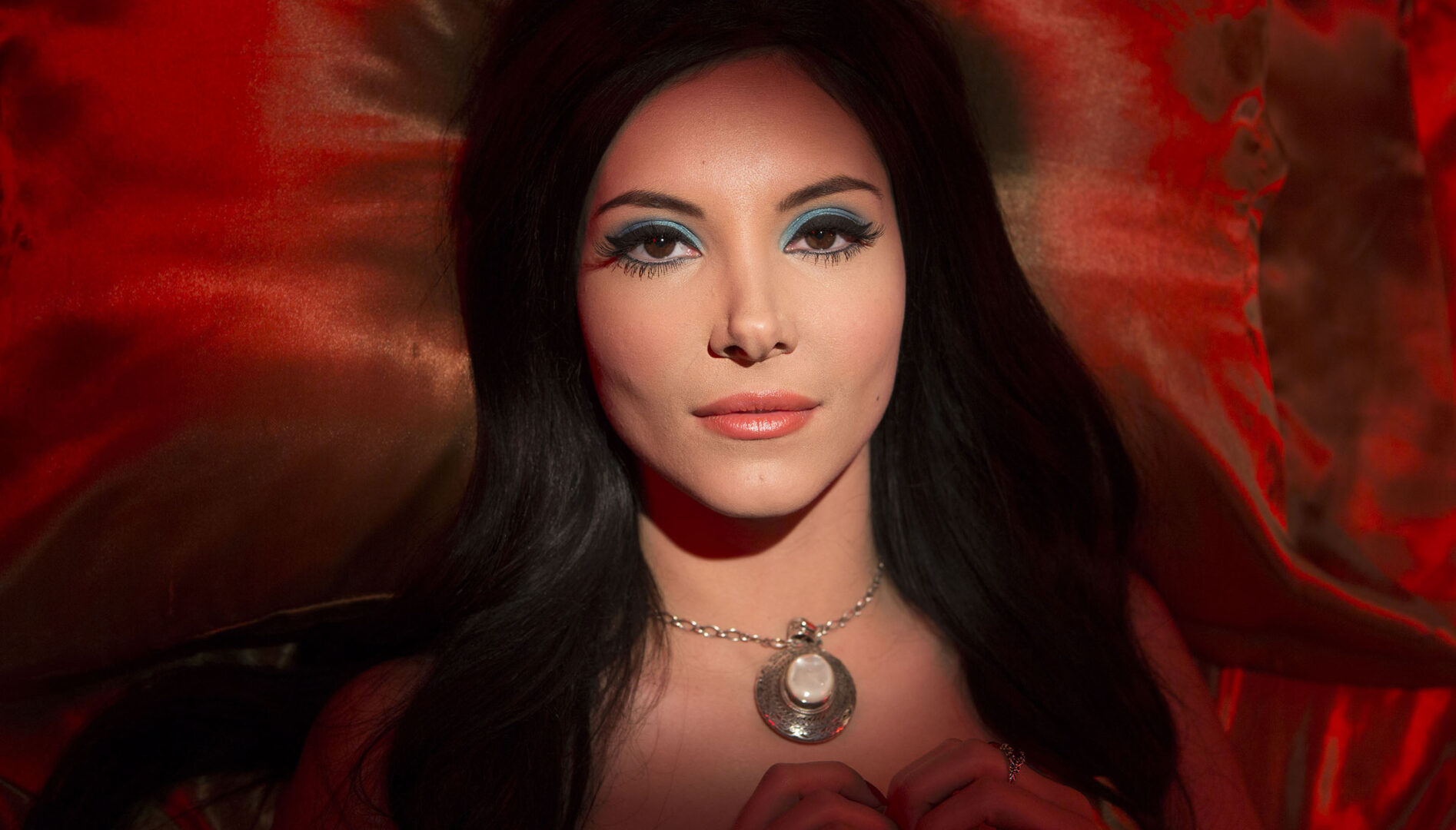 Close up photo of a woman with long brown hair, blue eye shadow, and a pendant necklace lying on a red velvet background