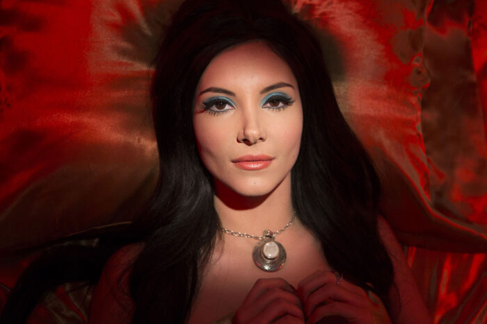 Close up photo of a woman with long brown hair, blue eye shadow, and a pendant necklace lying on a red velvet background