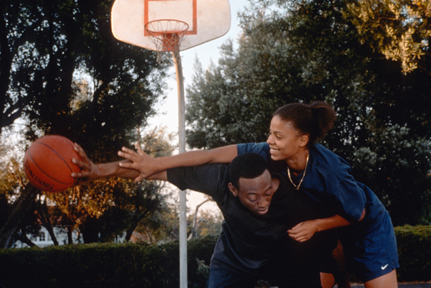 A young Black couple playing basketball on an outdoor court. She is jumping on his back trying to grab the ball.