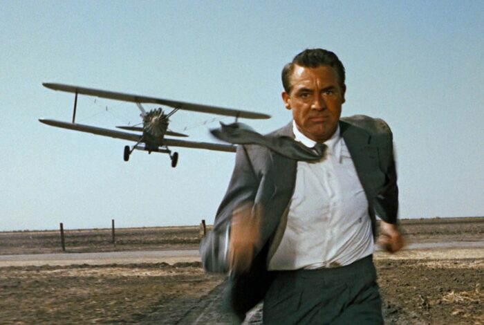 A man in a grey suit running in a field with a plane flying behind him