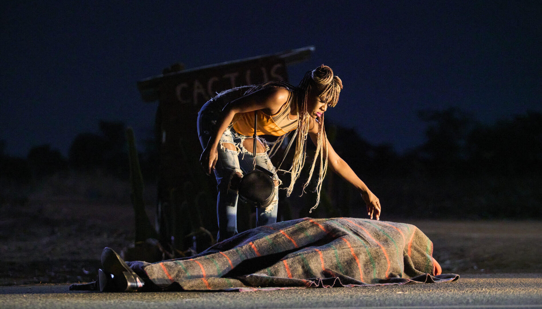 A Black woman with long dreadlocks leaving over a figure lying on the ground wrapped in a blanket