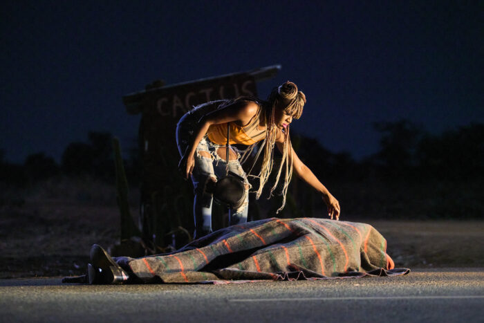 A Black woman with long dreadlocks leaving over a figure lying on the ground wrapped in a blanket