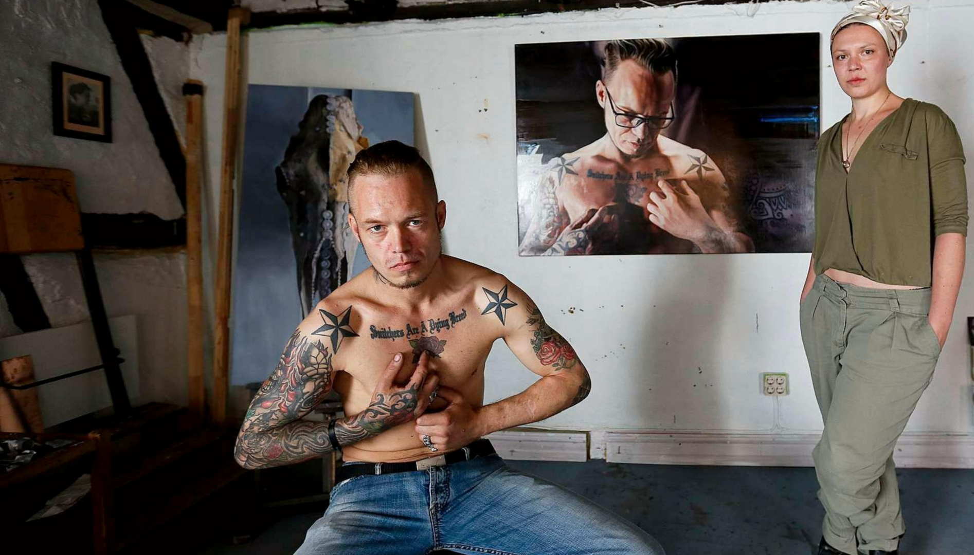 Photo of a woman standing in front of a painting of a man holding his arms up in a boxing pose. The subject of the painting, a topless tattooed man, is sitting in the middle of the studio.