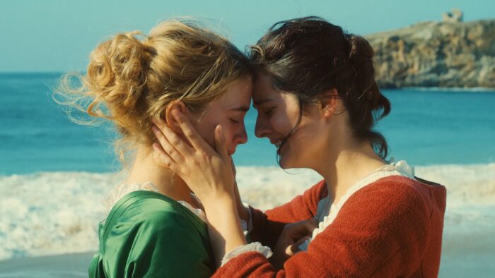 Photo of two women in front of the ocean holding their heads together. One is holding onto the other's face.