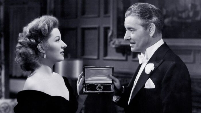 Black and white film still of a man and woman in formal dress holding a necklace in a jewelry box between them