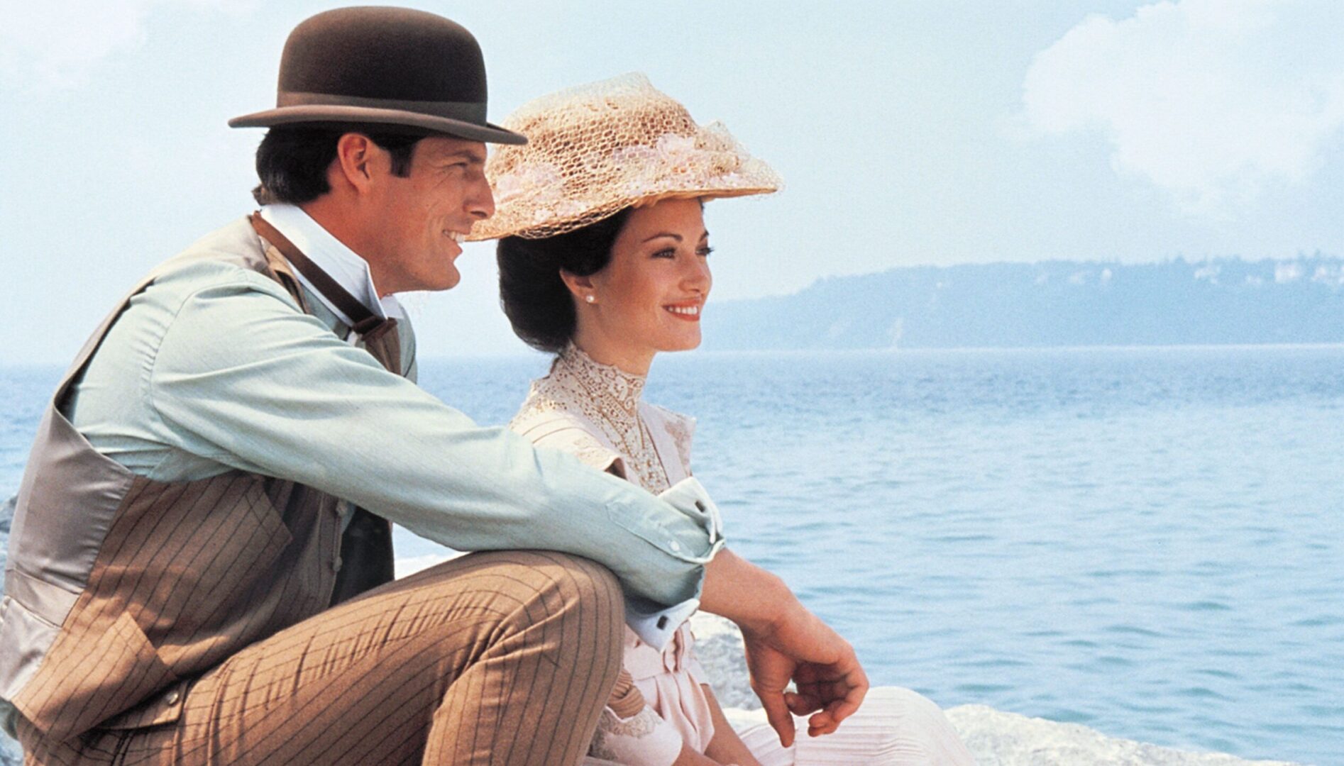 A man in a suit and a bowler hat and a woman in a fancy gown and hat sit next to a body of water, both looking to the side