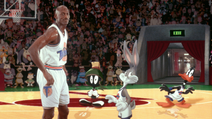 A tall Black man (Michael Jordan) standing on a basketball court with animated Looney Tunes figures–Bugs Bunny, Daffy Duck, and Marvin the Martian