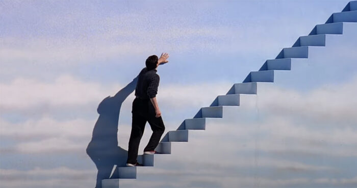 A man walks up a long staircase against a wall painted like a blue sky with clouds