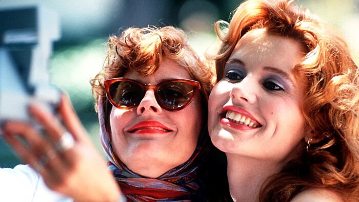 Two women with curly hair, one in sunglasses, smiling into a camera