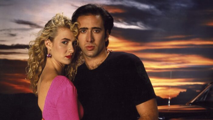 A woman with long, curly blond hair and a pink shirt and a man in a black t-shirt standing in front of a sunset