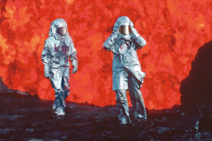 Two figures in silver fire-proof suits walk in front of a background of lava