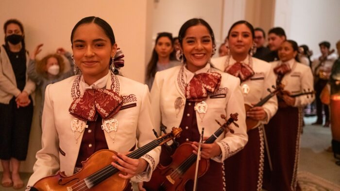 A group of young woman with their hair in ponytails, wearing white bolero jackets, maroon wide bow ties, and silver jewelry. They are holding violins and violin bows.