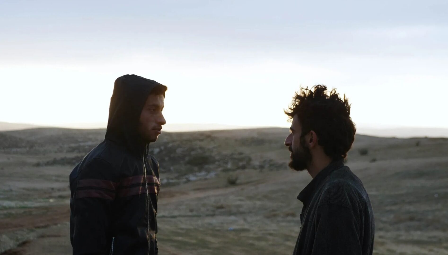A photo of two men facing each other, in a desert-like landscape with low light