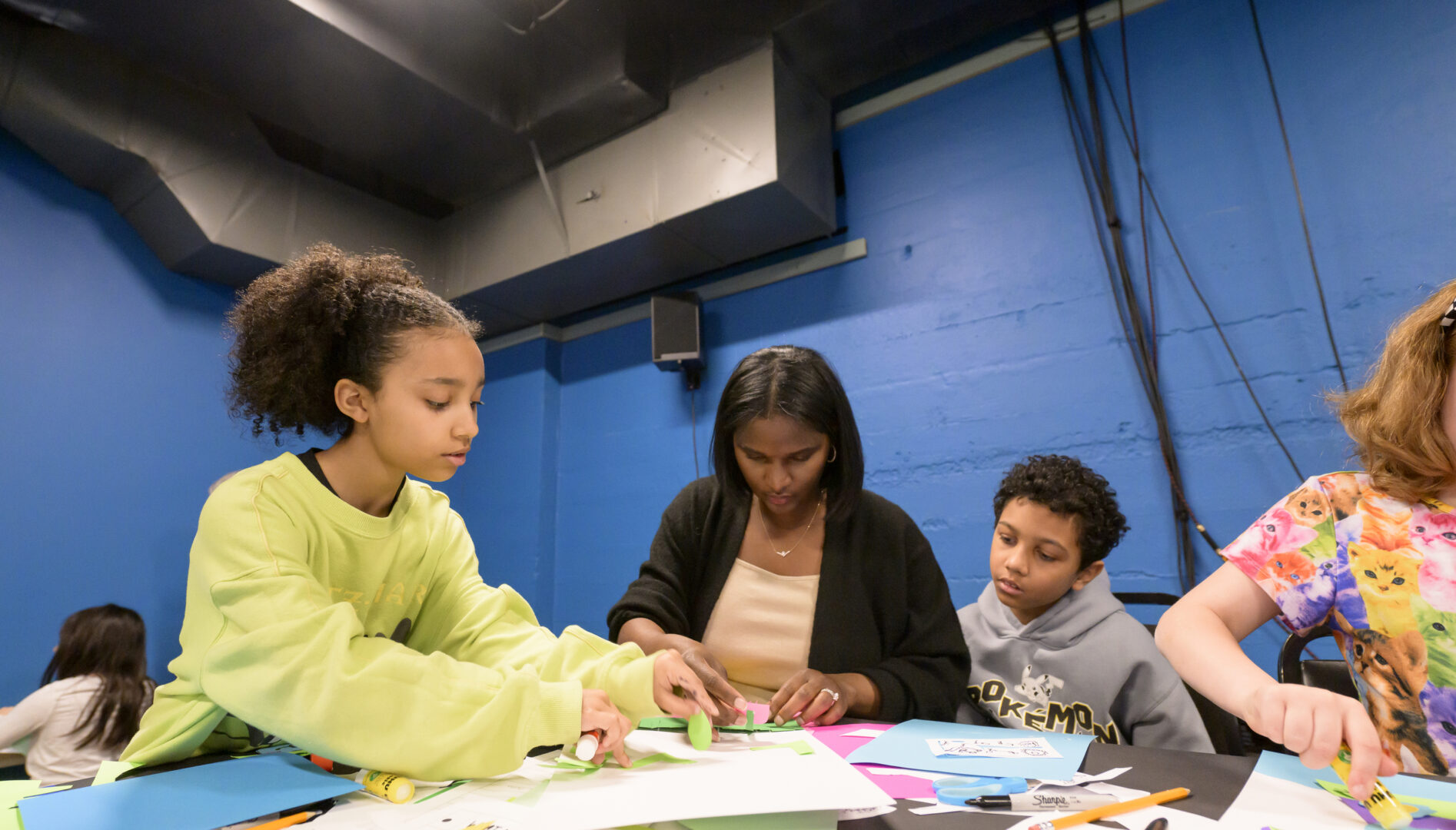 A mother sits between her daughter and son, all looking down at a table of paper, glue sticks, and other craft materials.