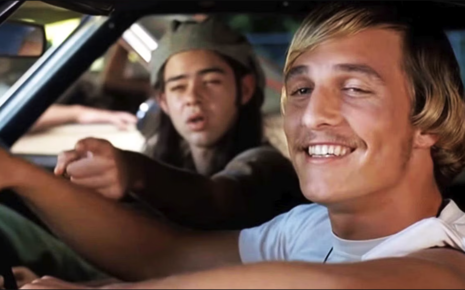 Film still of two young men in the front seat of a car. In the passenger's seat, a teenager with long hair and a hat on backwards. In the driver's seat, a man with blonde hair, a mustache, and a white tee shirt with stoned-looking eyes and a smile on his face.