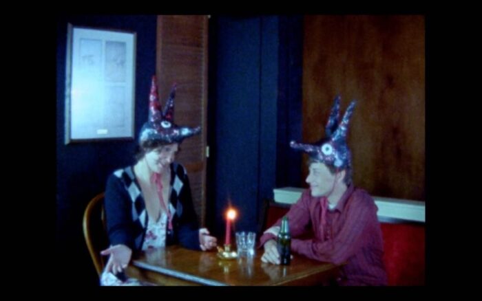 A film still of two people sitting at a table, with animal masks on top of their heads. There's a lit candle, a cup, and a beer on the table.