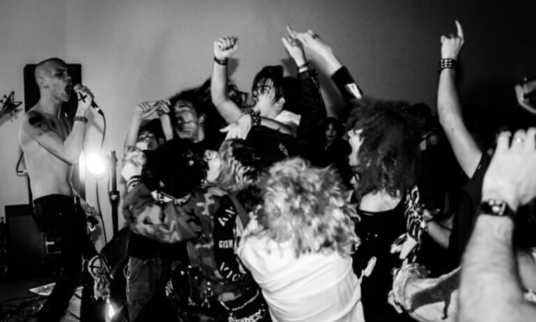 Black and white photo of a crowd of people dancing at a punk show. A singer with a shaved head is singing in the lefthand side of the photo.