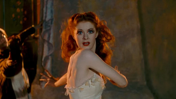 Film still of a woman with long red hair and a sleeveless off-white dress spinning towards the camera with an alarmed look on her face.