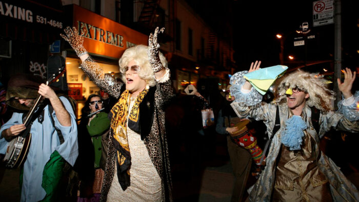 Photo of a group of people, some playing instruments, some in drag, walking down a city street at night