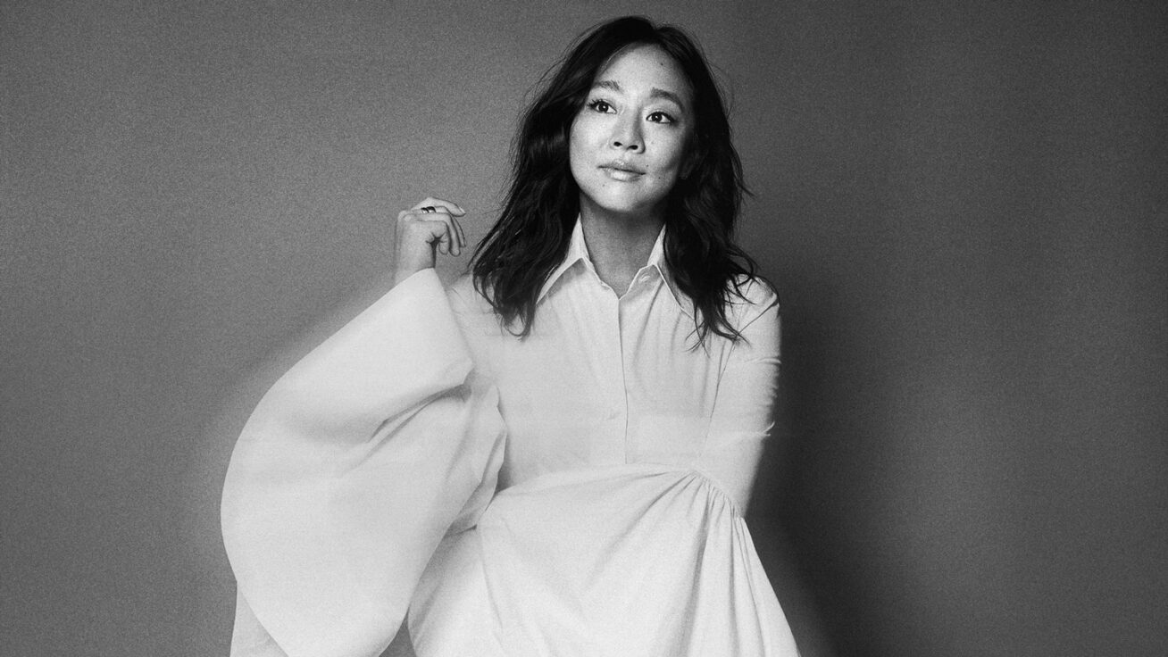 A black and white photo of a woman with shoulder length dark hair, wearing a white button-down dress with huge sleeves.