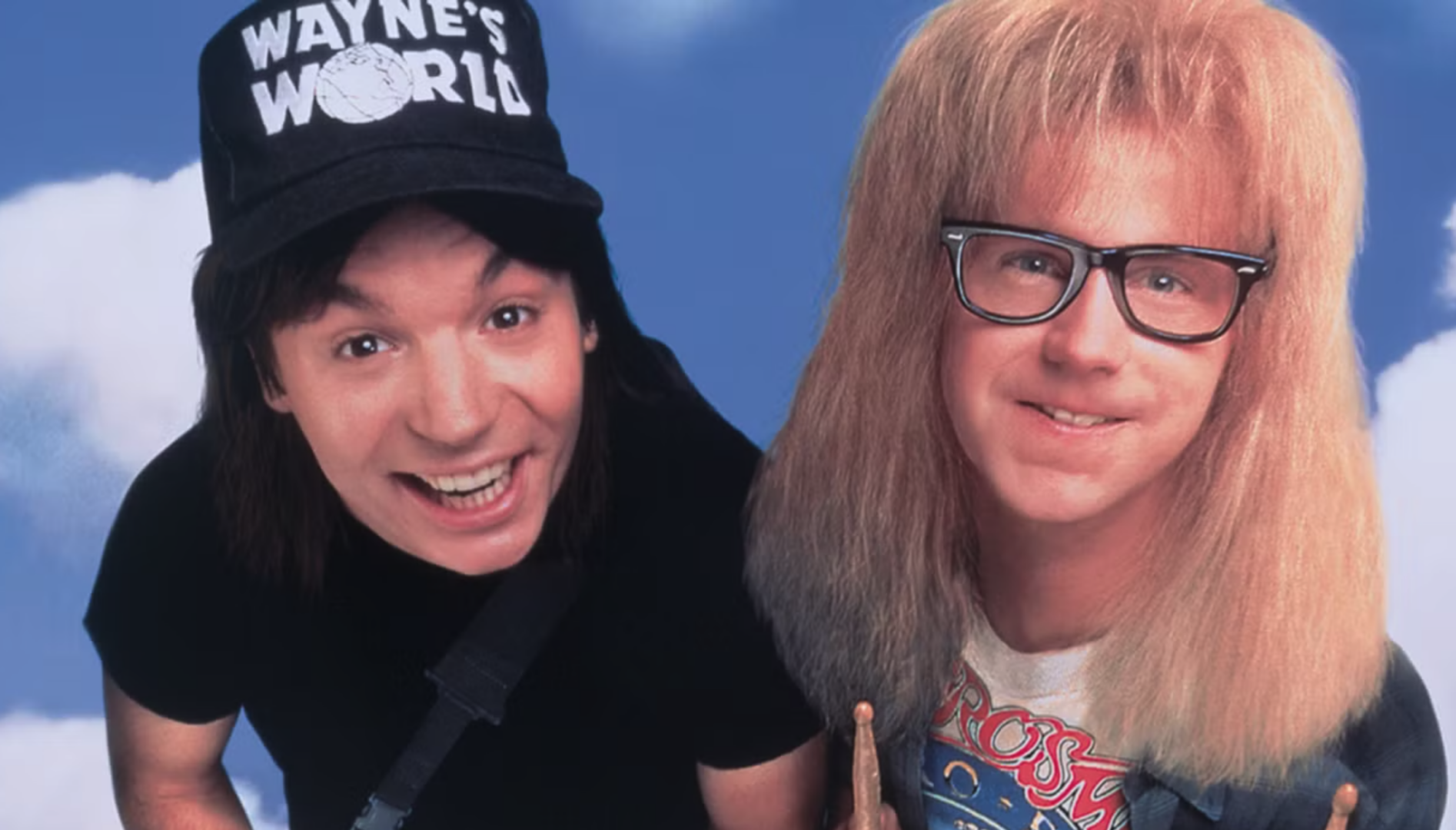 A film still of two smiling men standing in front of a blue sky. One has long brown hair and a black hat that says "Wayne's World"; the other has long blond hair and black eyeglasses.