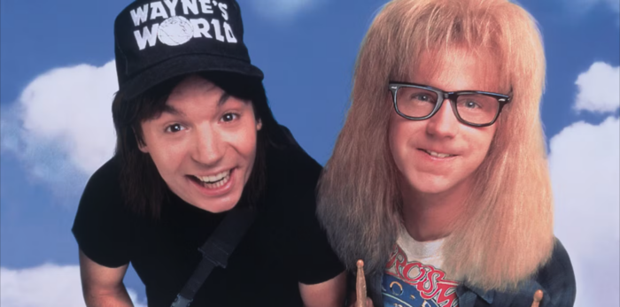 A film still of two smiling men standing in front of a blue sky. One has long brown hair and a black hat that says "Wayne's World"; the other has long blond hair and black eyeglasses.