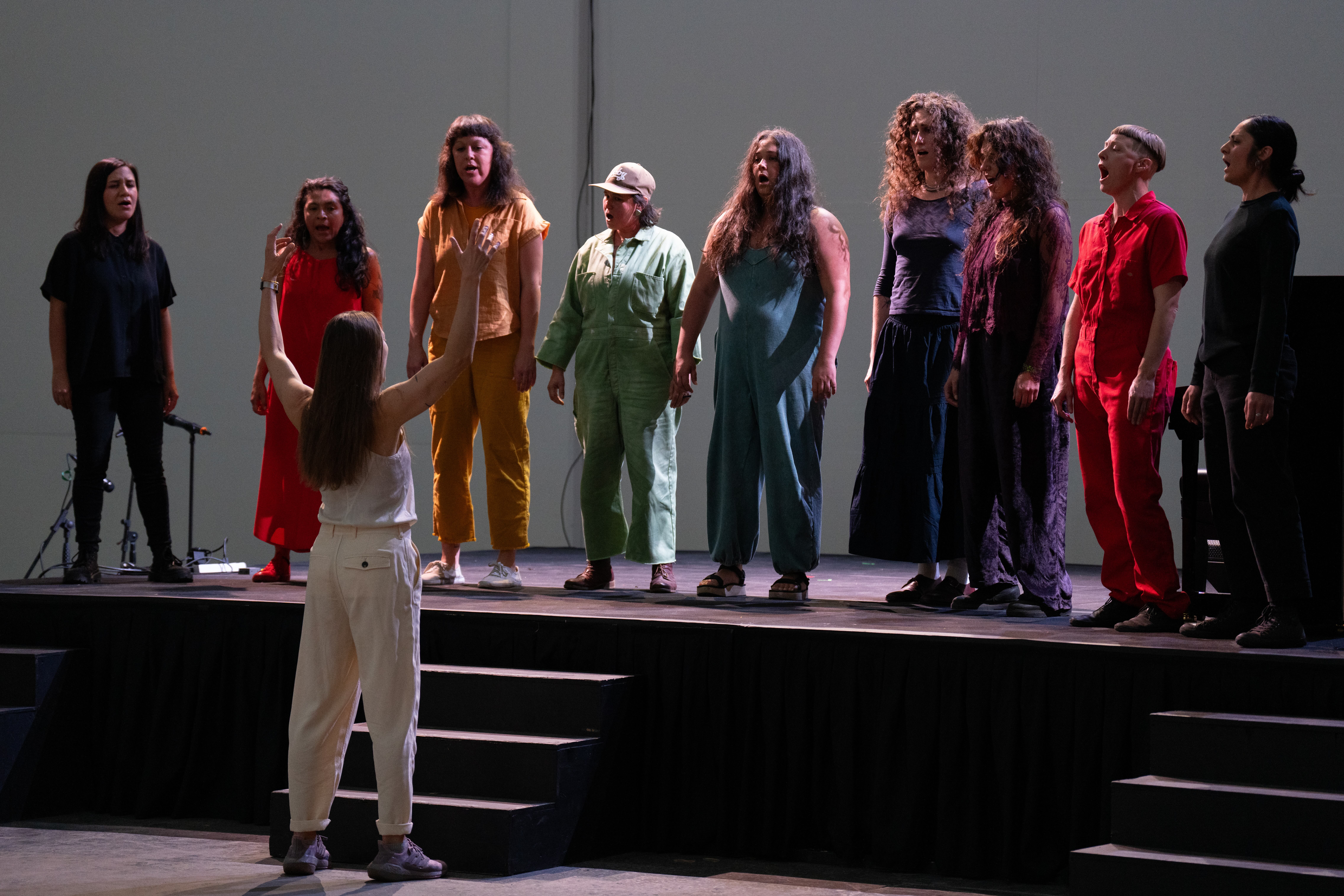A group of people in different colored jumpsuits singing on a stage. A person on the floor is conducting them.